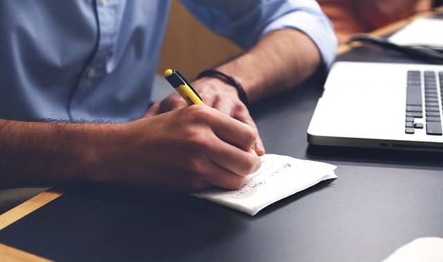 Photo of a man, sitting in front of a laptop, a pencil in one hand, writing something on a notebook. Maintaining a TO DO list for programming is easy to start.