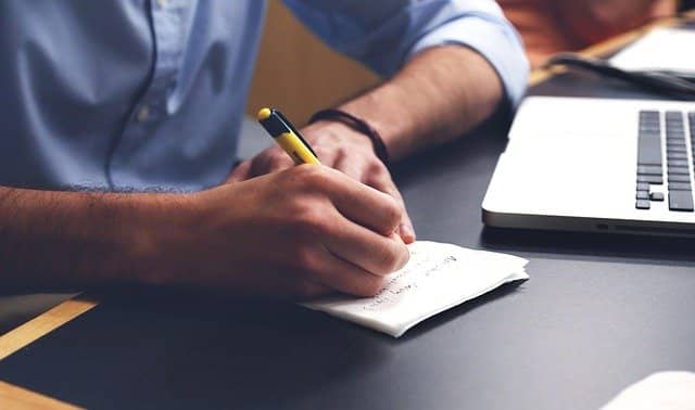 Photo of a man taking notes on a paper notebook next to his computer. Taking digital notes when remote pair coaching is even better than traditional paper notes in co-localized pairing. We can use this to walk coachees through a more organized, predictable, productive, and sustainable way of programming.